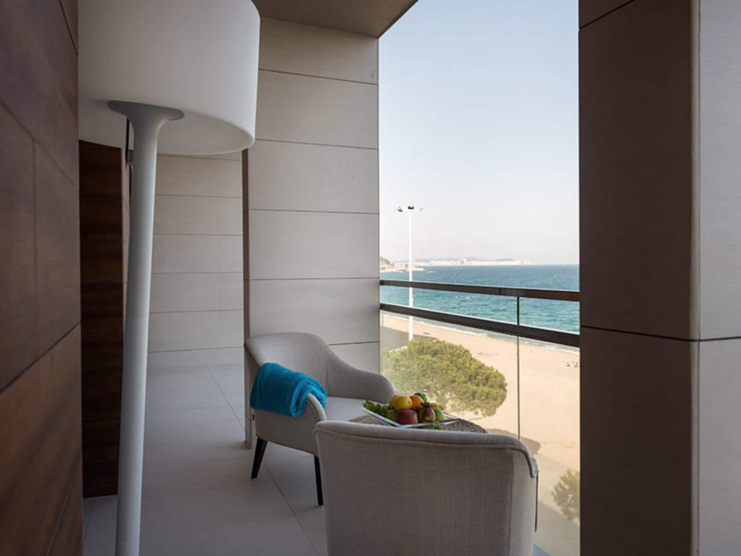 A balcony with a view of the sea, a chair, a table with fruit, and a modern lamp.