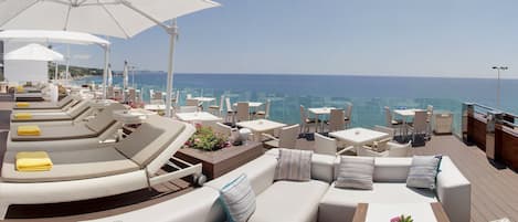 A sunlit outdoor seating area with lounge chairs and umbrellas, overlooking the sea.