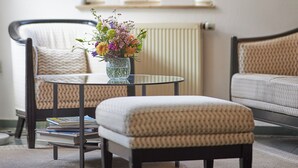 A modern living room with a round glass table, two patterned armchairs, a floor lamp, and a vase with flowers. - Nassauer Hof Limburg (Limburg an der Lahn)