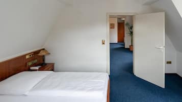 Habitación de hotel con cama, mesita de noche con lámpara, radio y puerta que da a un pasillo con planta en maceta.