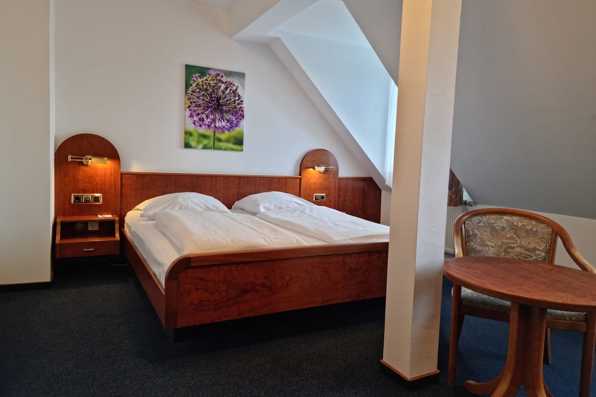 A hotel room with a wooden bed, two bedside tables with lamps, a chair, and a painting of flowers on the wall.