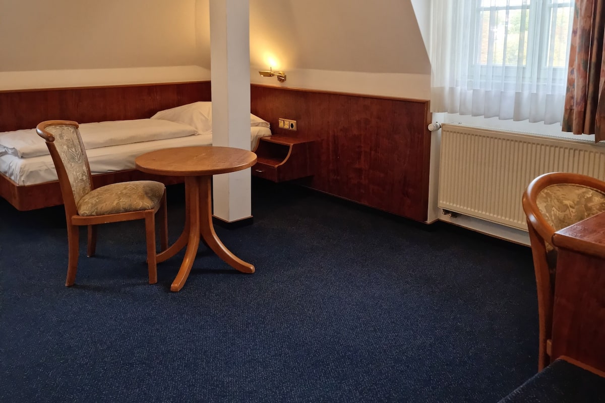 A room with a bed, a wooden table, a chair, and a window with curtains.