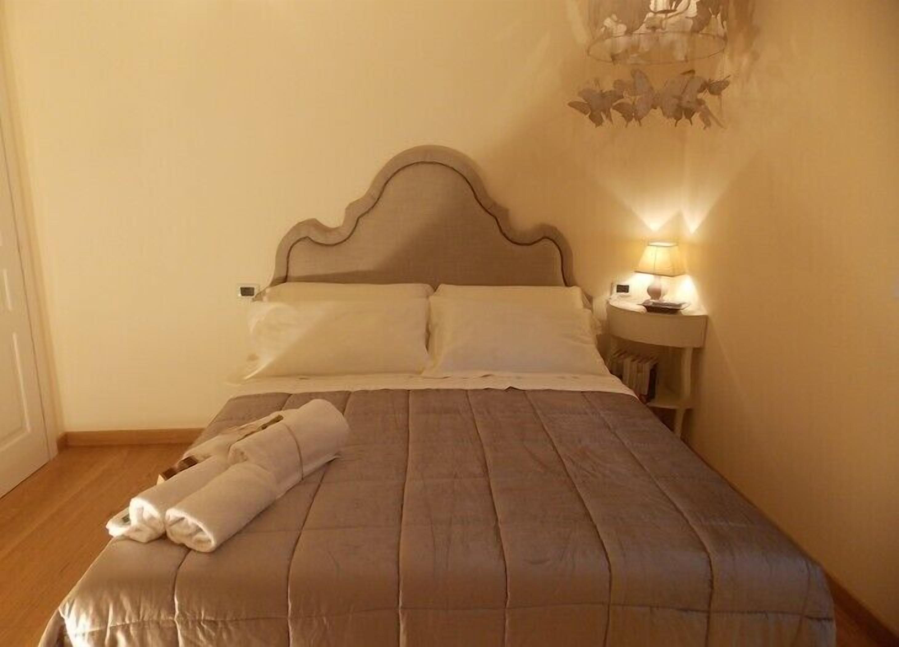 Uma cama bem arrumada com cabeceira, mesa de cabeceira com abajur e luminária fixada na parede.