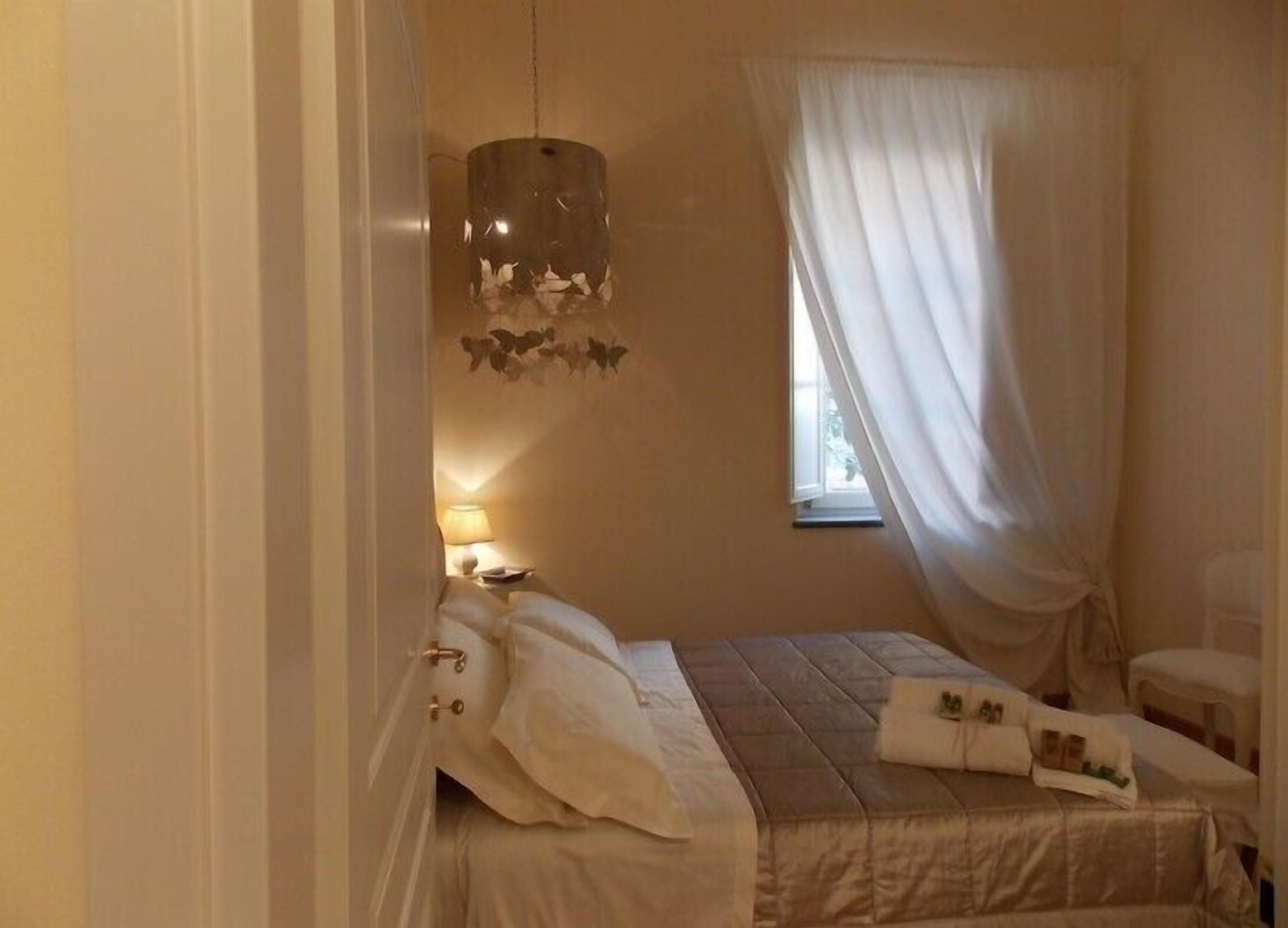 A bedroom with a bed, a hanging light fixture, a window with sheer curtains, and a nightstand with a lamp.