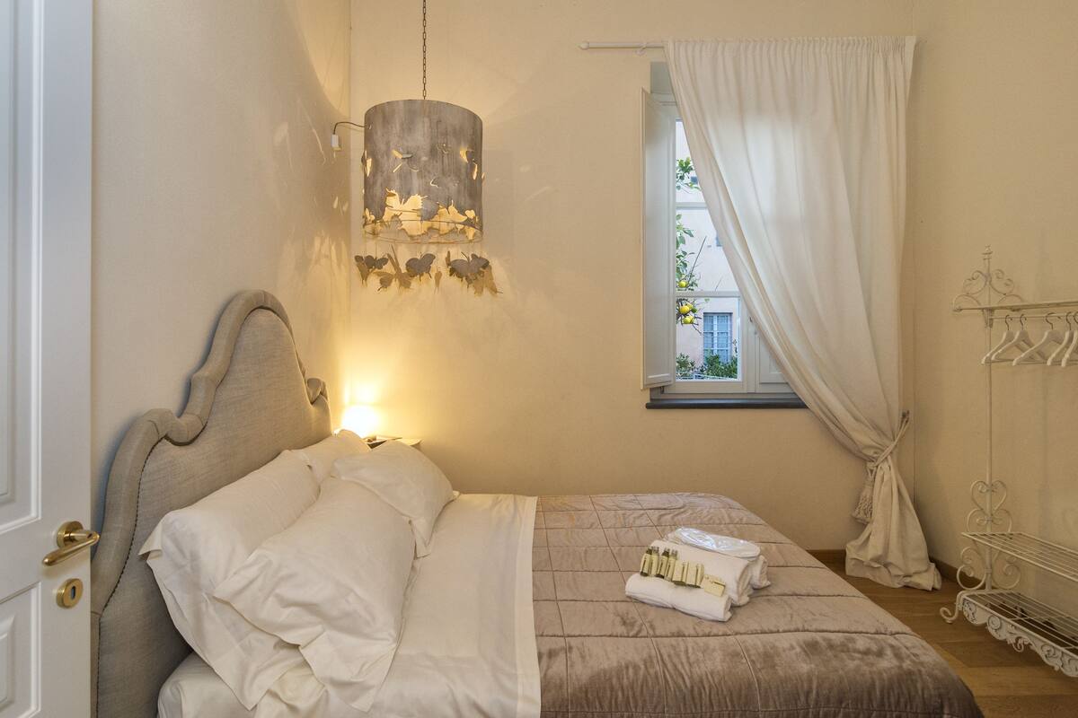 Un lit bien fait, avec du linge de lit blanc, un luminaire suspendu orné de feuilles, une fenêtre avec des rideaux et un portant métallique avec des cintres.