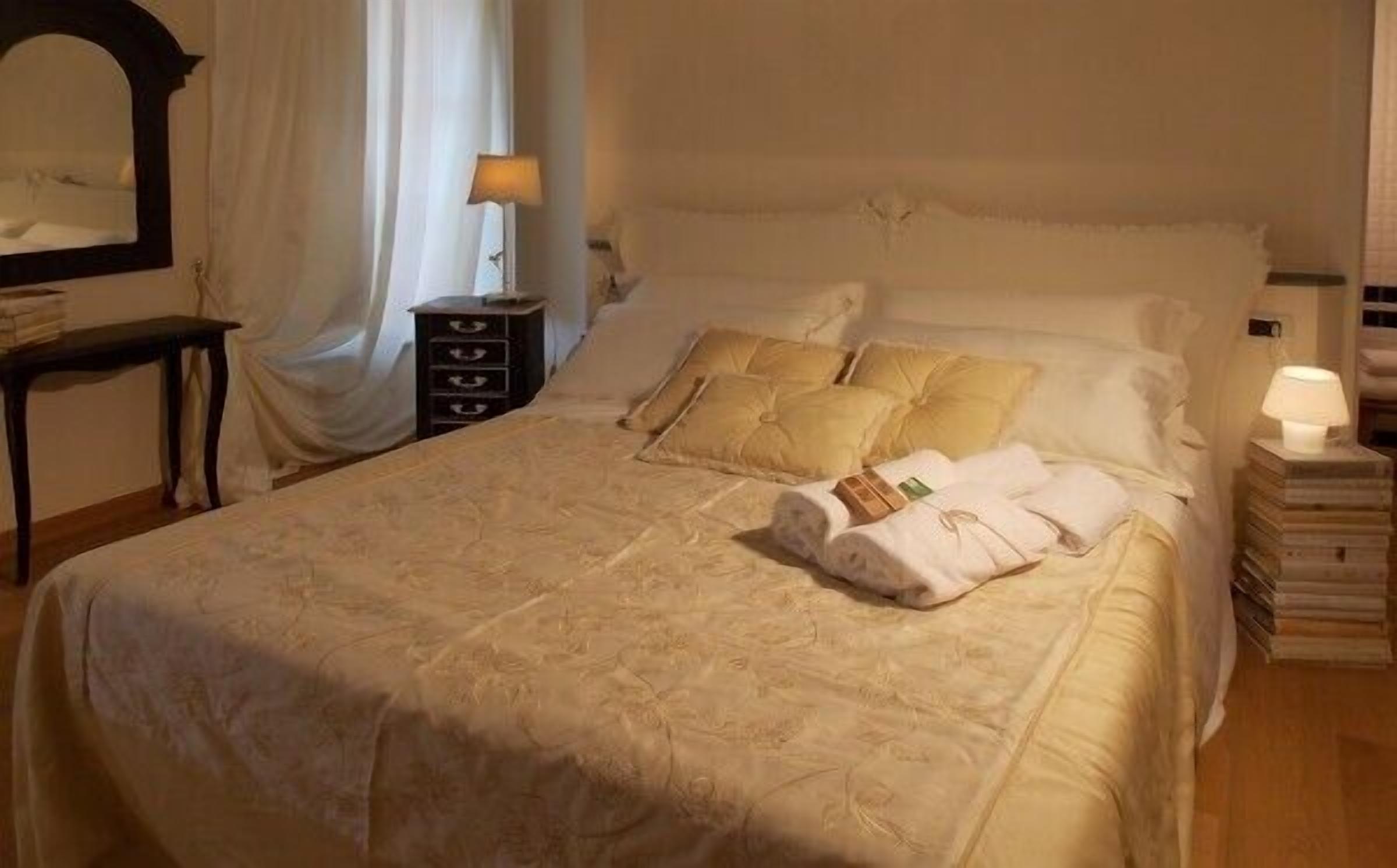 Un letto rifatto con cura, con piumone, cuscini e un asciugamano arrotolato. È presente un comodino con una lampada e una pila di libri.