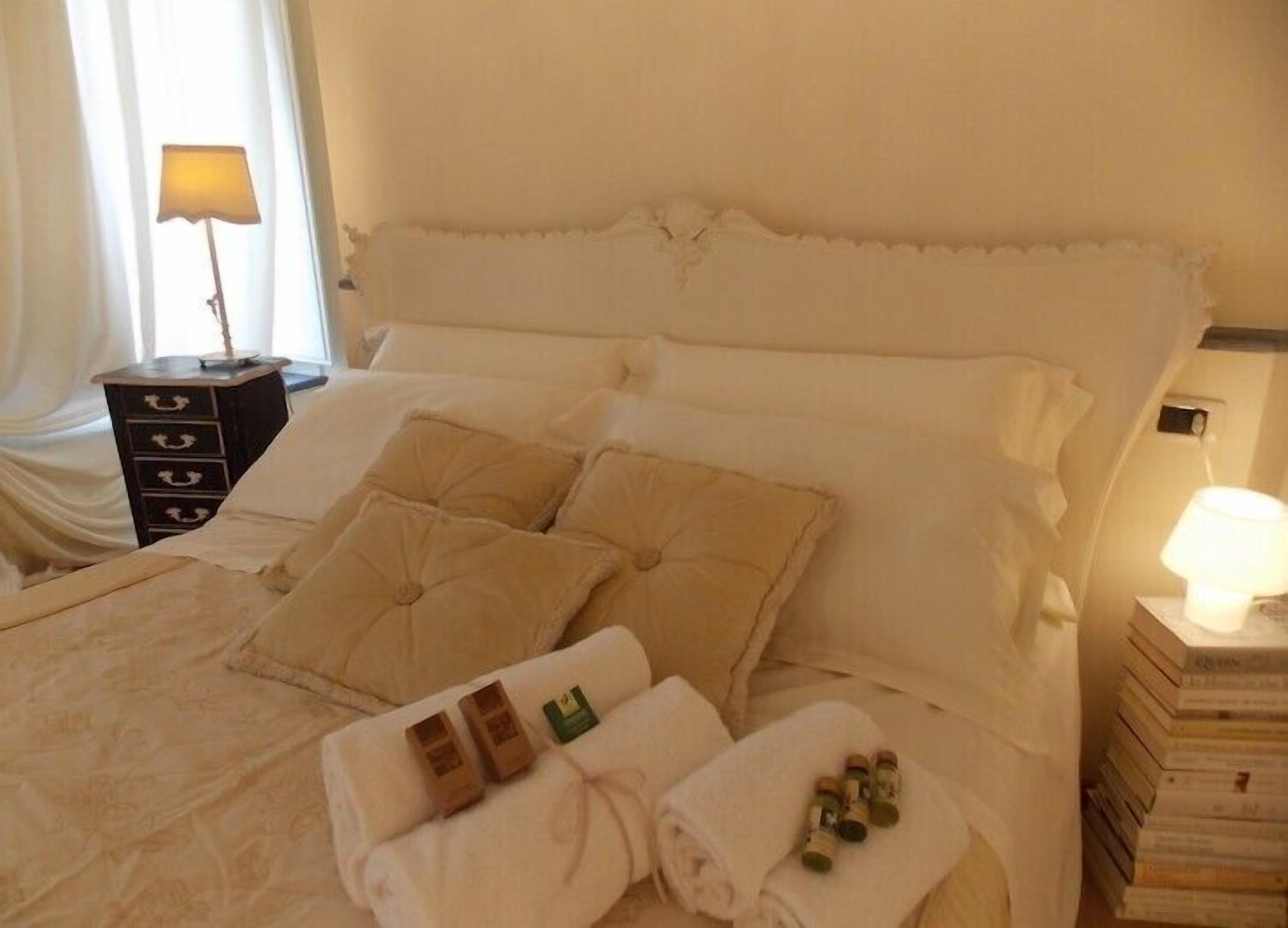 Un letto rifatto con cuscini, un comodino con una lampada e una pila di libri accanto al letto.