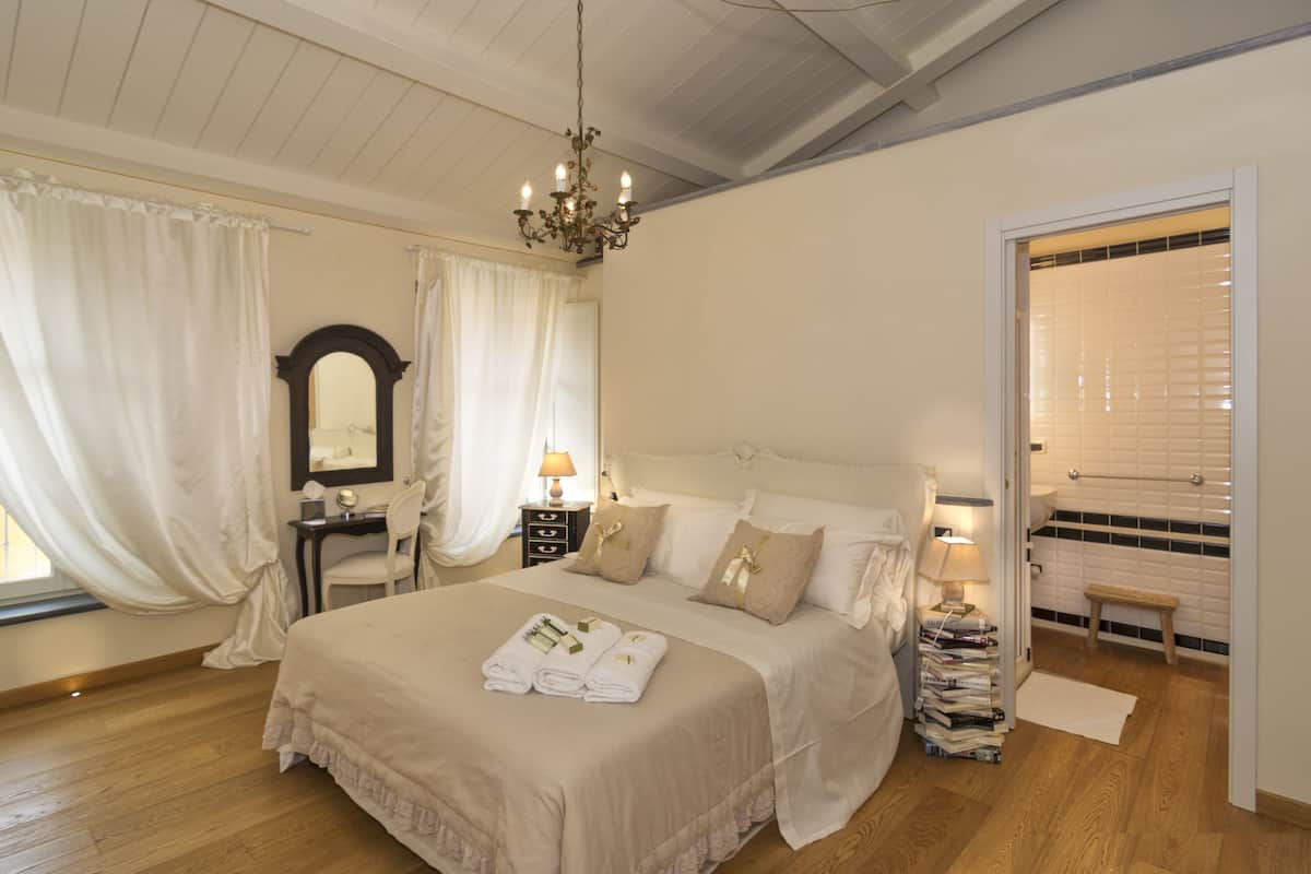Une chambre à coucher avec un grand lit, un lustre, une table de chevet avec une lampe, un miroir et une fenêtre avec des rideaux.