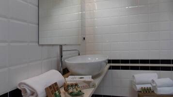 Un baño con azulejos blancos, un lavamanos grande, un espejo y un banco de madera con toallas y artículos de aseo.