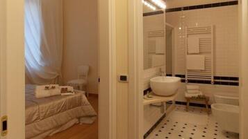 Een slaapkamer met een bed, een stoel en een nachtkastje met een lamp. De badkamer heeft een wastafel, spiegel en toilet.