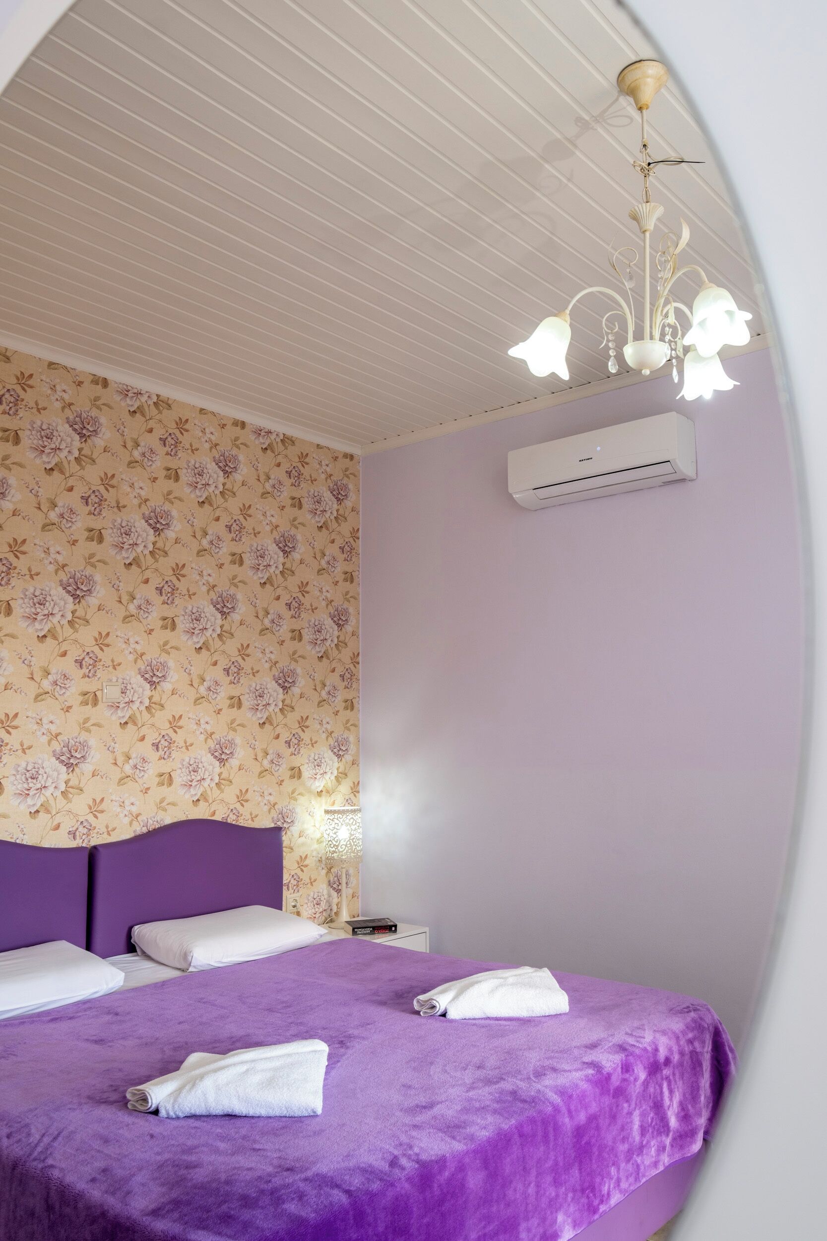 A bedroom with two beds, a floral wallpaper, a purple bedspread, a white air conditioning unit, and a chandelier.