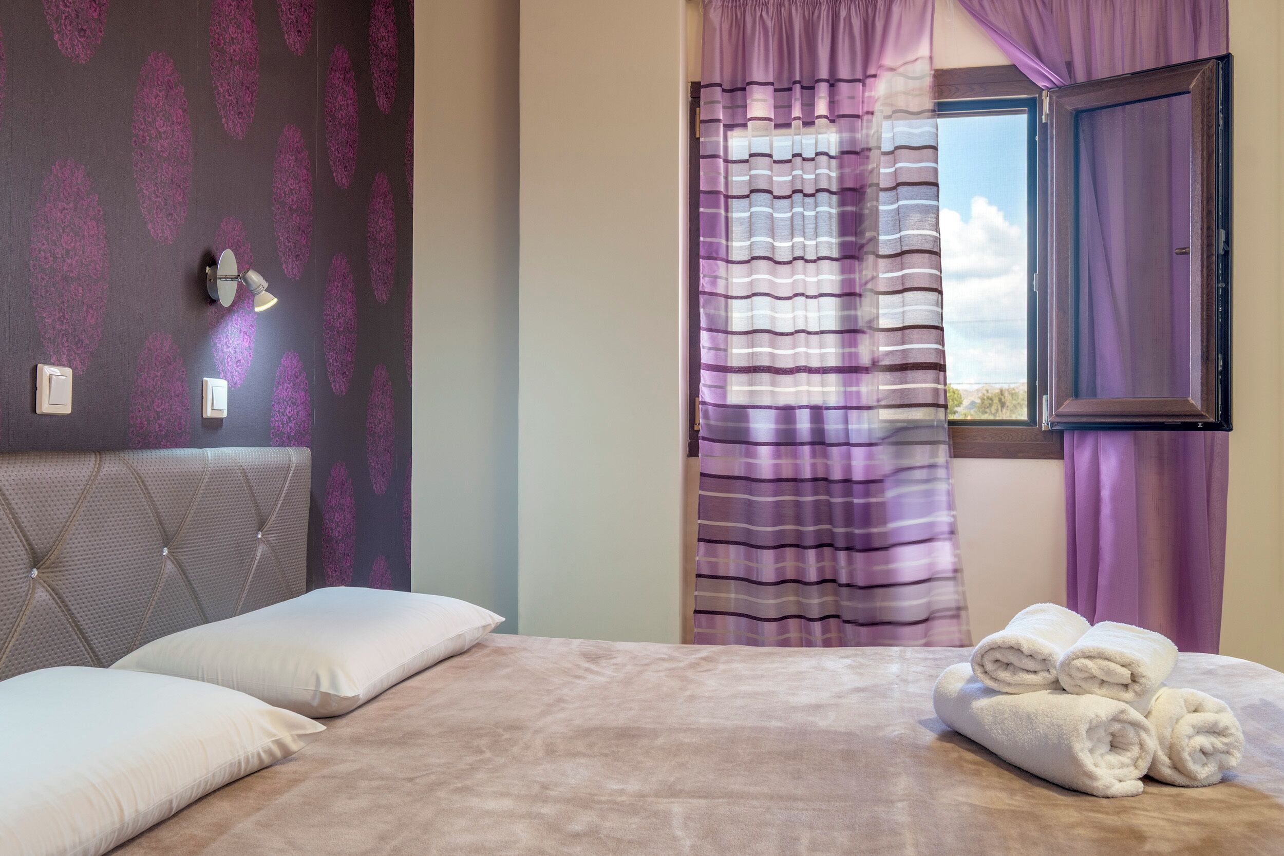 Une chambre à coucher avec un lit, des oreillers, une fenêtre avec des rideaux et un mur orné d’un motif.