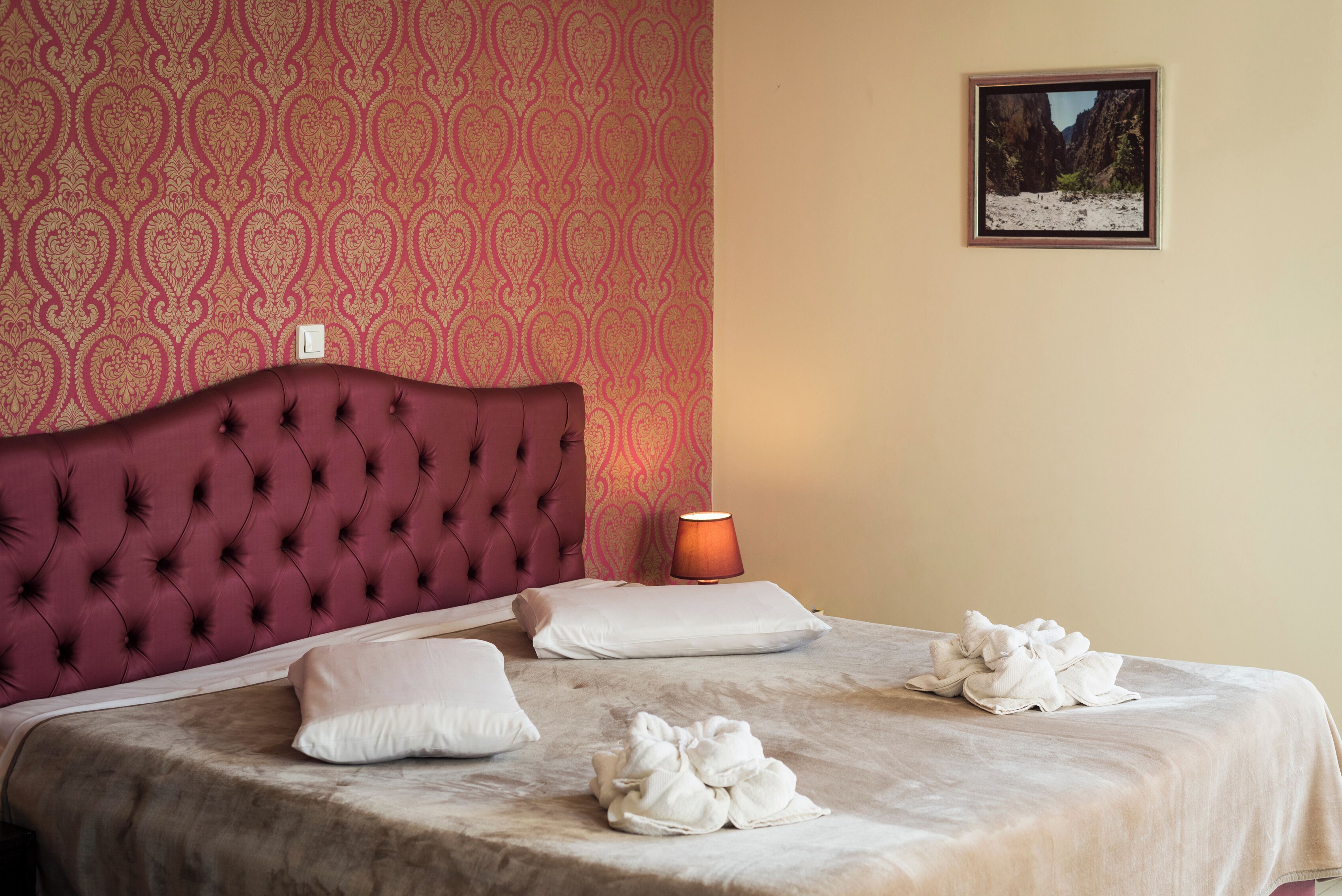 Una cama bien hecha con cabecera roja, almohadas blancas y toallas sobre un edredón color beige.