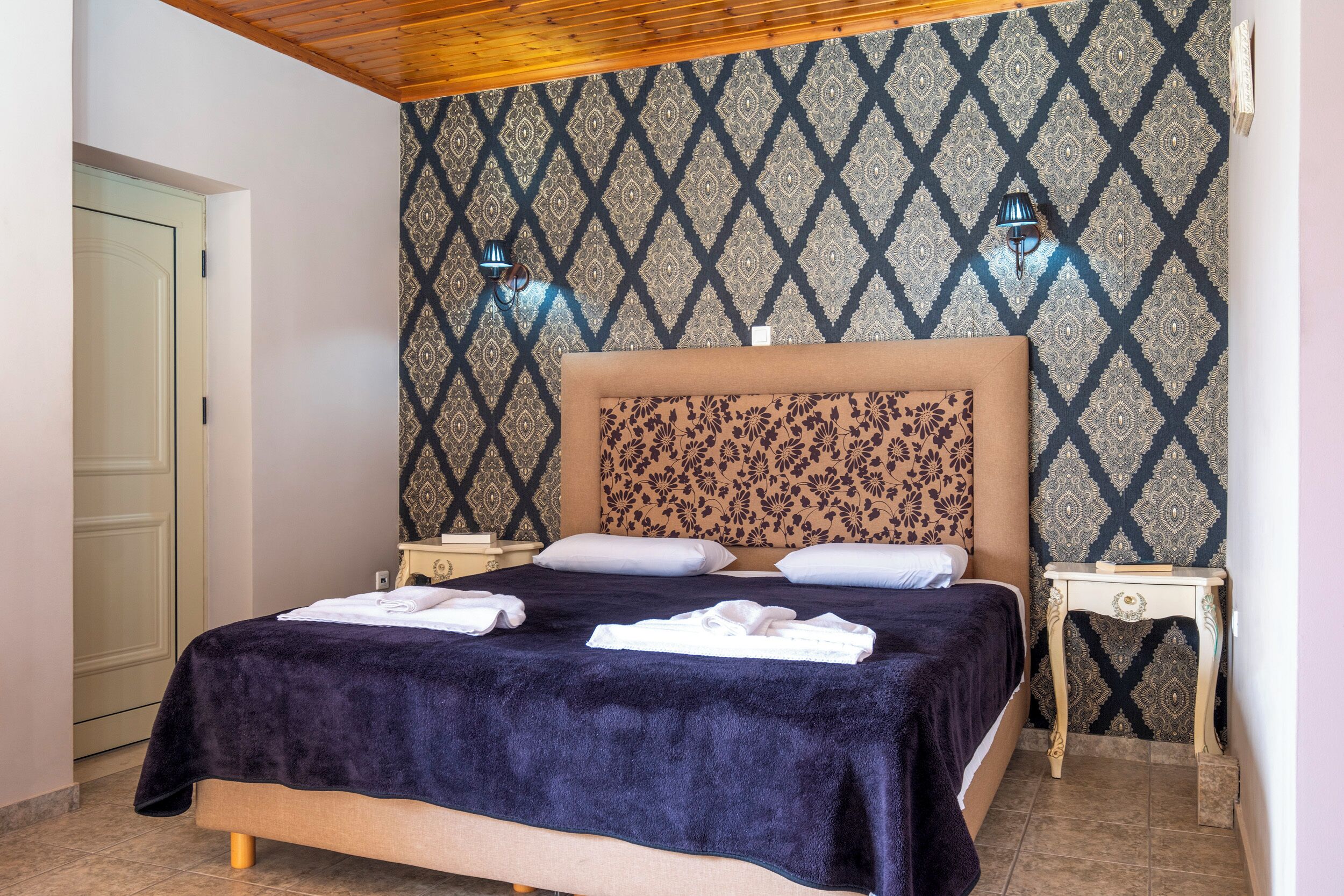 A neatly made bed with a dark blue blanket, two bedside tables with lamps, and patterned wallpaper.