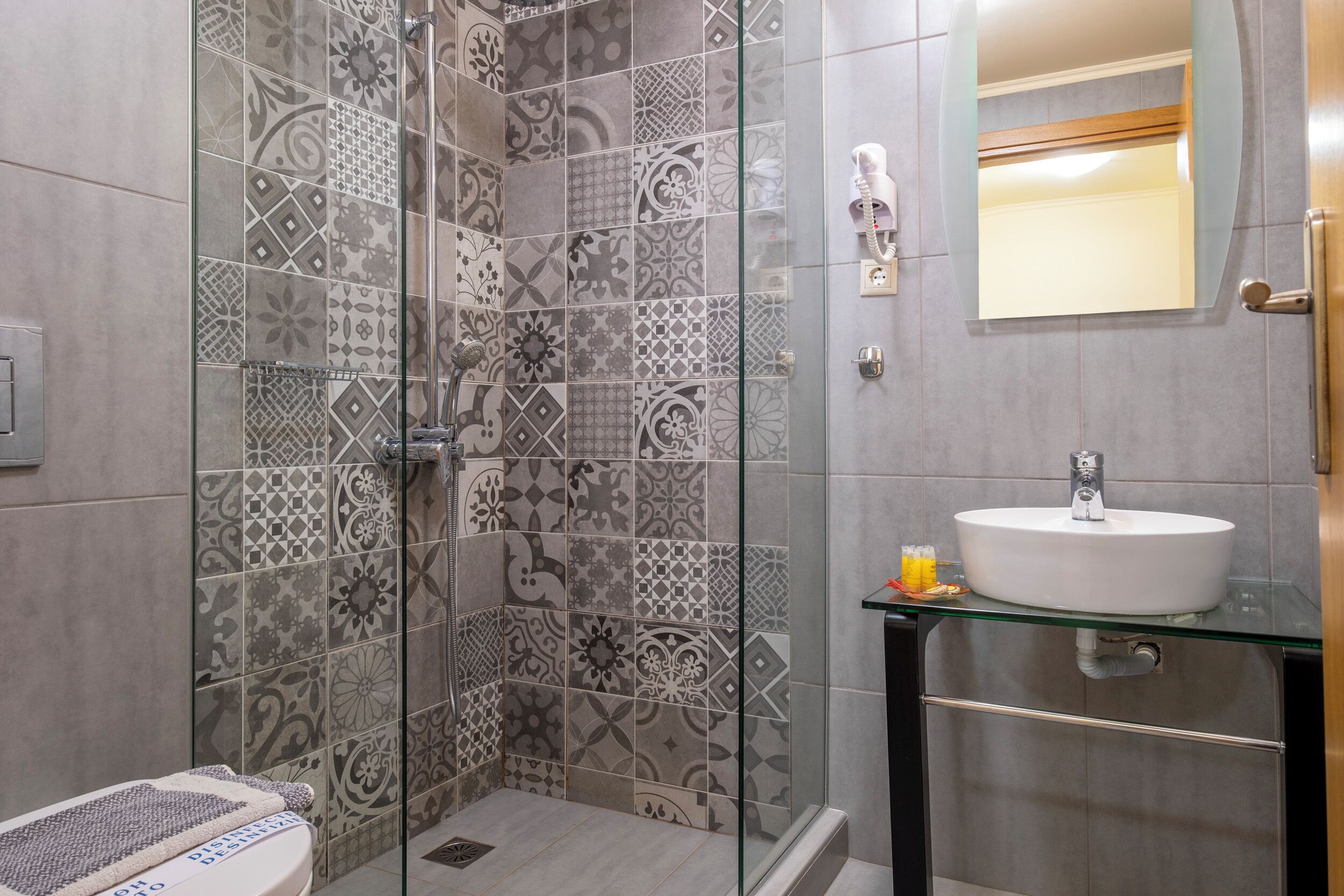 A bathroom with patterned tiles, a glass shower enclosure, a sink with a glass countertop, and a mirror above the sink.