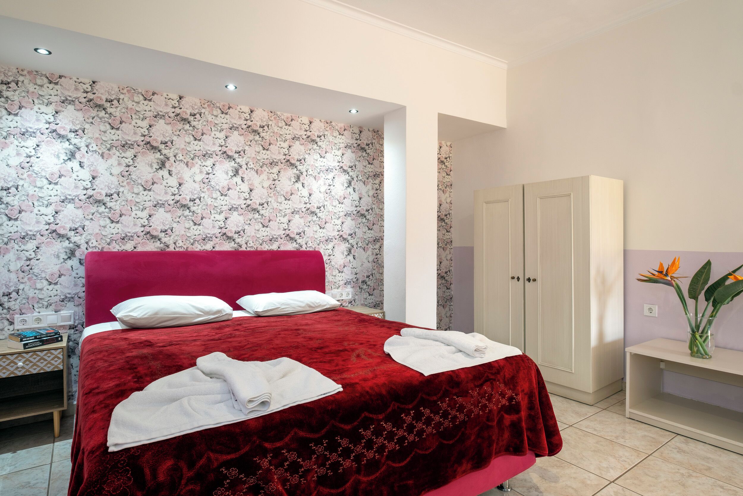 Habitación de hotel con una colcha roja, almohadas blancas, una mesita de noche con libros y un armario.