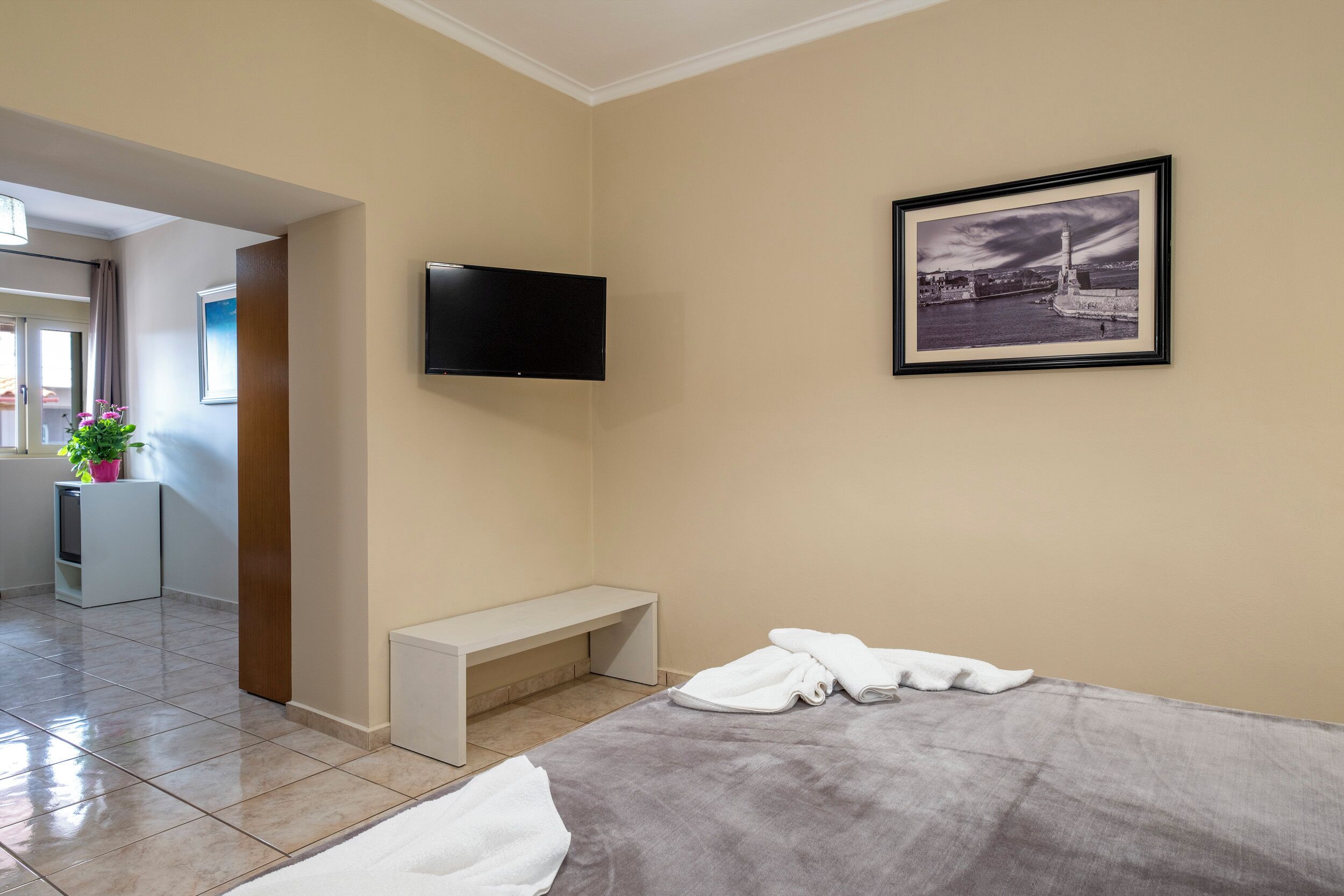 A hotel room with a bed, a bench, a flat-screen TV, a framed picture, and a small kitchenette.