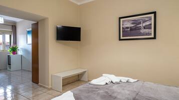 A hotel room with a bed, a bench, a flat-screen TV, a framed picture, and a small kitchenette.