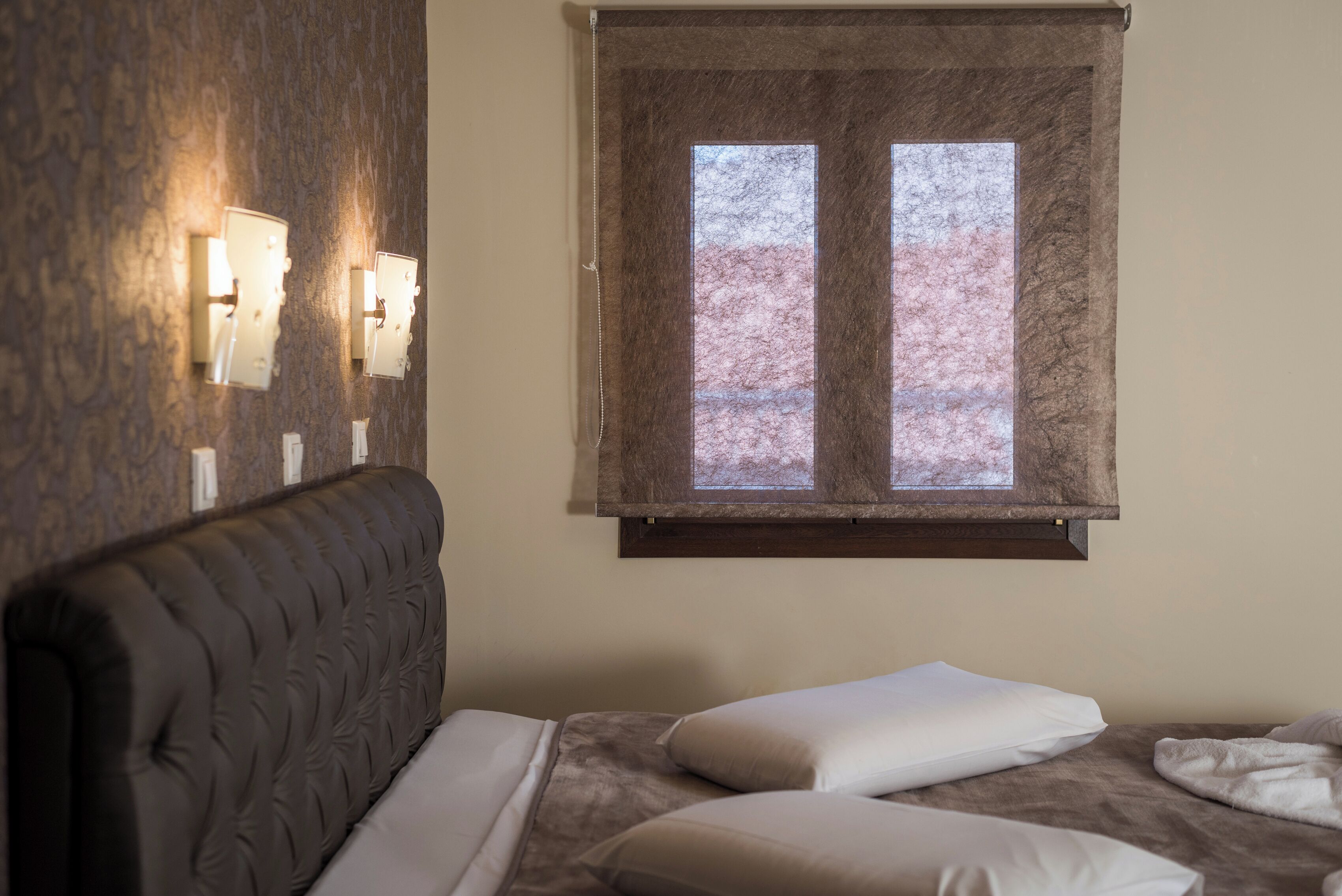 A hotel room with a bed, a headboard, two wall-mounted light fixtures, and a window with blinds.
