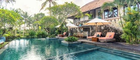 A pool area with lounge chairs and umbrellas, surrounded by tropical vegetation and a stone building.