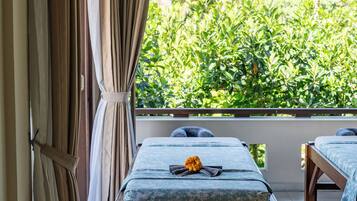 Relax in a serene room with a view of lush greenery, featuring a massage table, nightstand, and vase with flowers.
