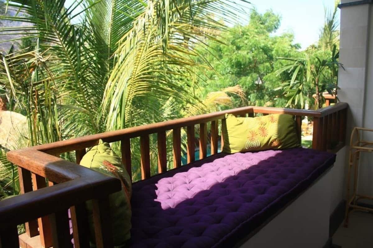A balcony with a wooden railing, a cushioned bench, and a view of tropical foliage.