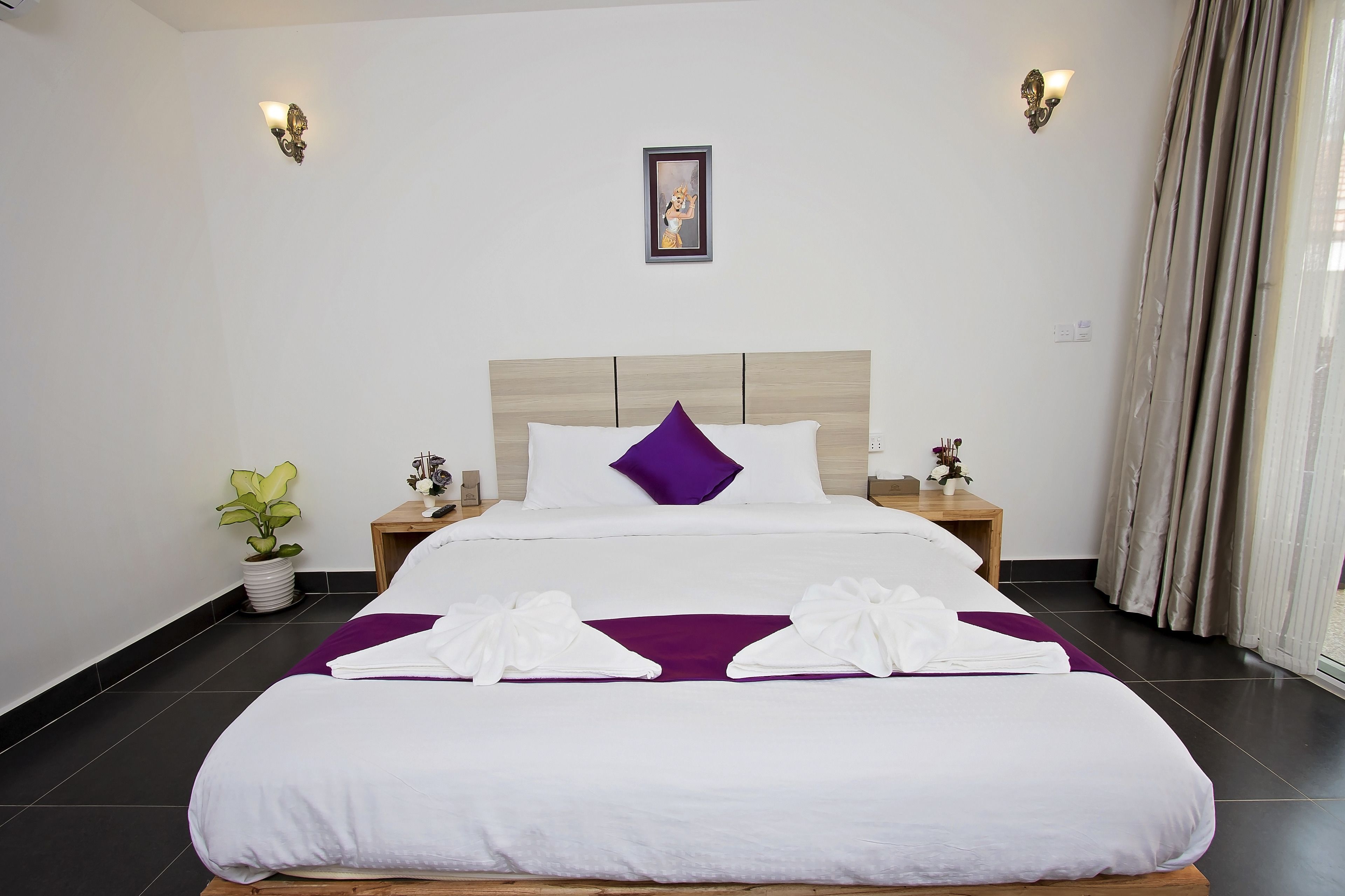 A neatly made bed with white and purple bedding, two bedside tables with lamps, a potted plant, and a framed picture on the wall.