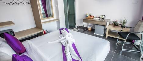Una habitación de hotel con una cama bien hecha, un escritorio con una computadora, una silla, una mesita de noche con artículos de aseo y un televisor en la pared.