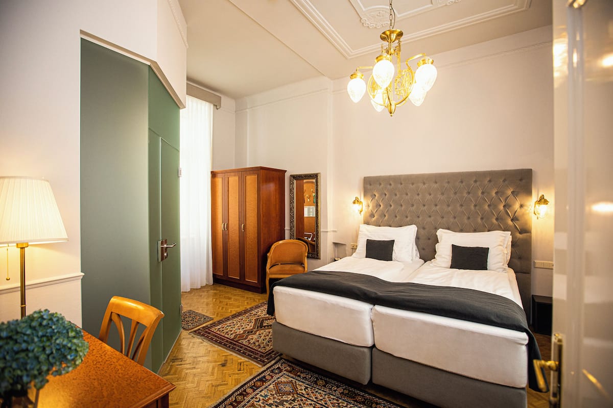 A hotel room with a large bed, a wooden wardrobe, a dining table with chairs, and a chandelier.