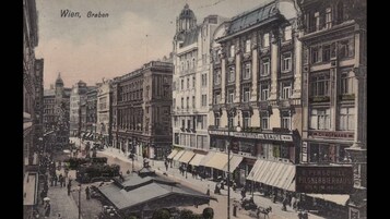 Cena de rua histórica em Viena, com edifícios elaborados, uma placa de destaque para 'C. Hofmann' e uma área de mercado com tendas.