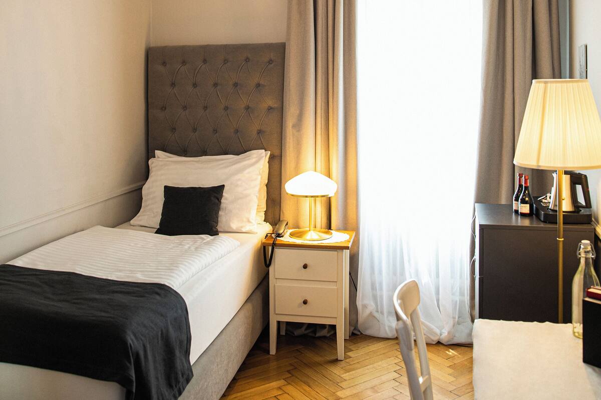 A hotel room with a bed, bedside table, lamp, chair, and a view of the outside through a window with curtains.