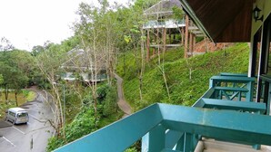 Une vue depuis un balcon donnant sur une route, avec un véhicule, une végétation luxuriante et des bâtiments traditionnels en arrière-plan.