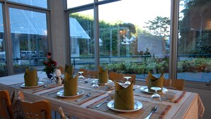 Une table à manger dressée avec des serviettes pliées, des assiettes, des couverts et des verres, offrant une vue sur la verdure et un bâtiment à l’extérieur.