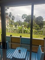 Vista a través de una ventana de una habitación con una mesa y bancos azules. En el exterior, se observa una cerca verde, un edificio blanco y abundante vegetación, incluyendo palmeras.