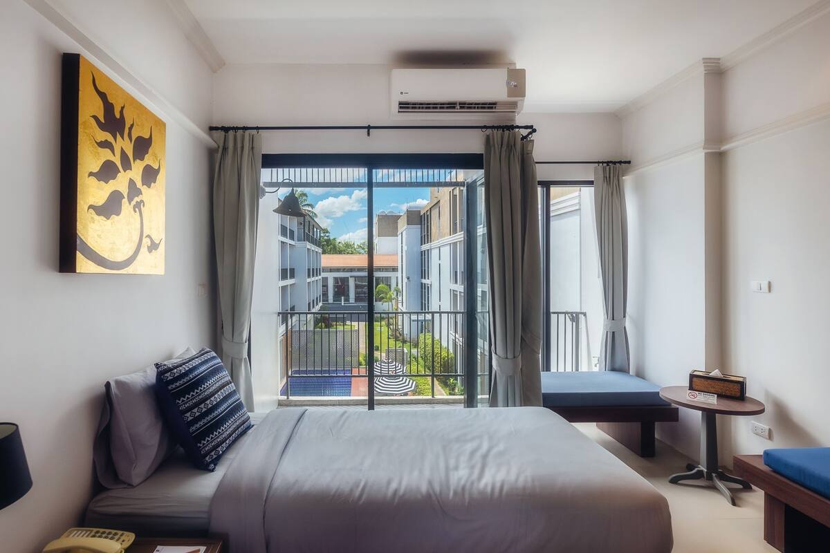 Habitación de hotel con una cama grande, un escritorio con teléfono y documentos, una mesita con caja de pañuelos y vistas a edificios desde un balcón con cortinas.