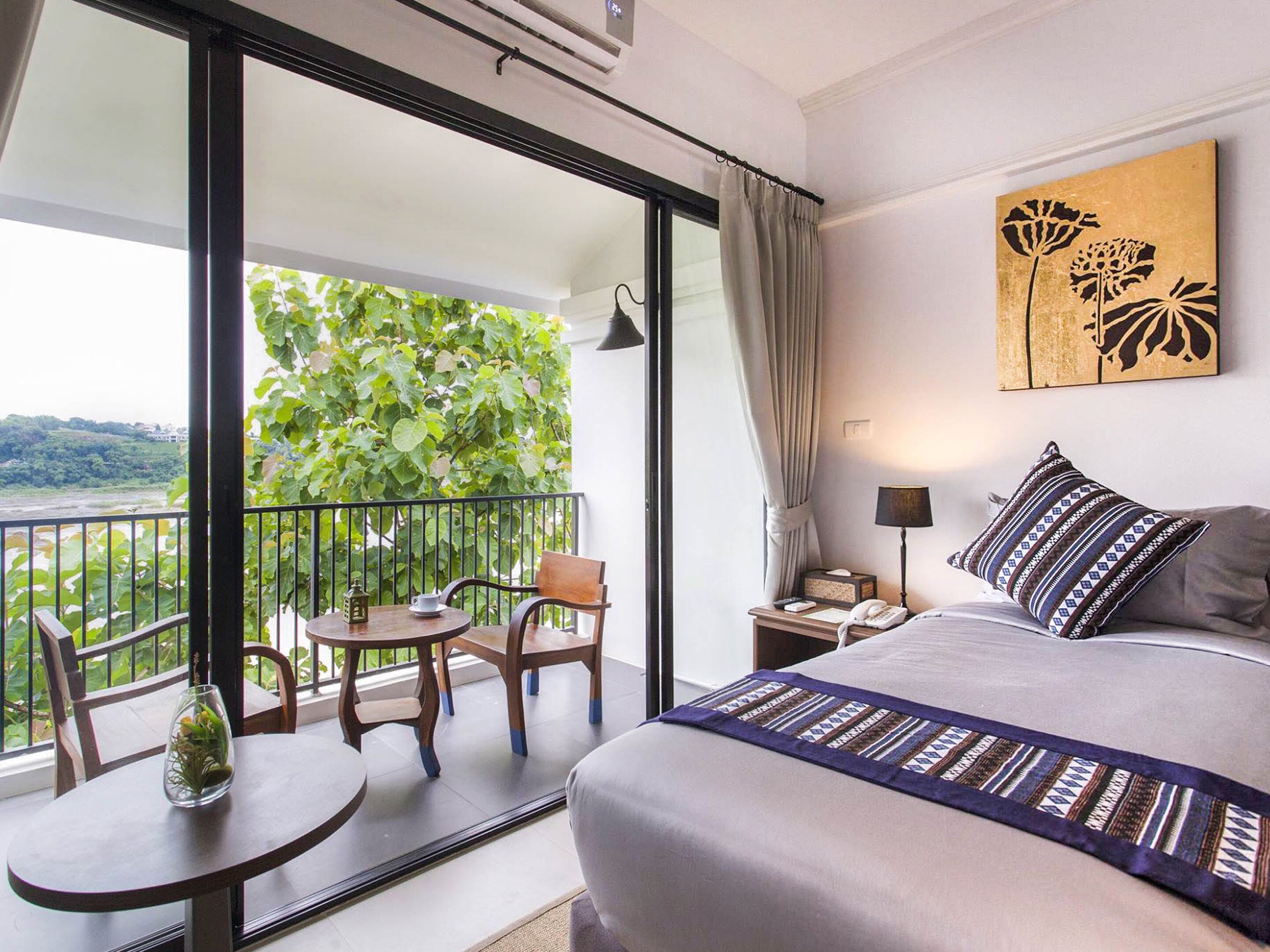 A hotel room with a balcony, a bed with a patterned bedspread, a nightstand with a lamp, and a view of greenery.