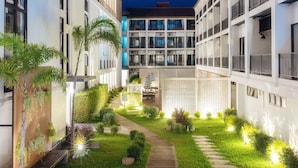 Une cour d’hôtel moderne avec un chemin central, entourée de verdure et de palmiers.