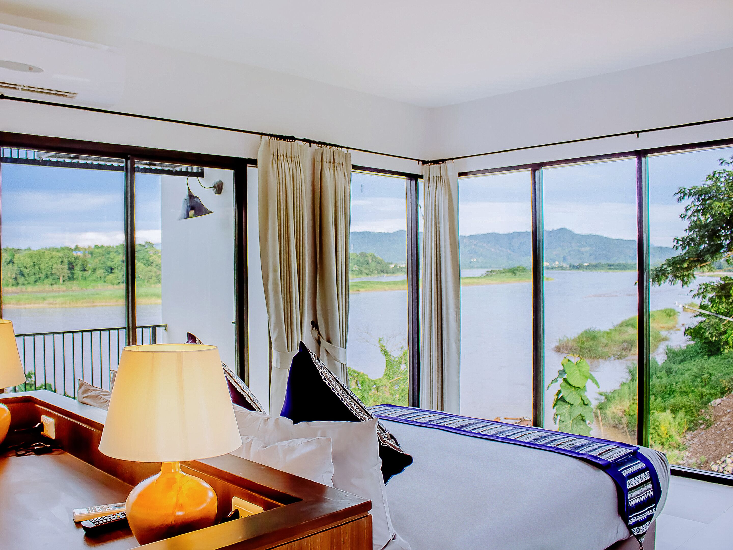 A hotel room with a large window offering a view of a lake and mountains. There is a bed with a white and blue bedspread, a bedside table with a lamp, and a remote control on the table.