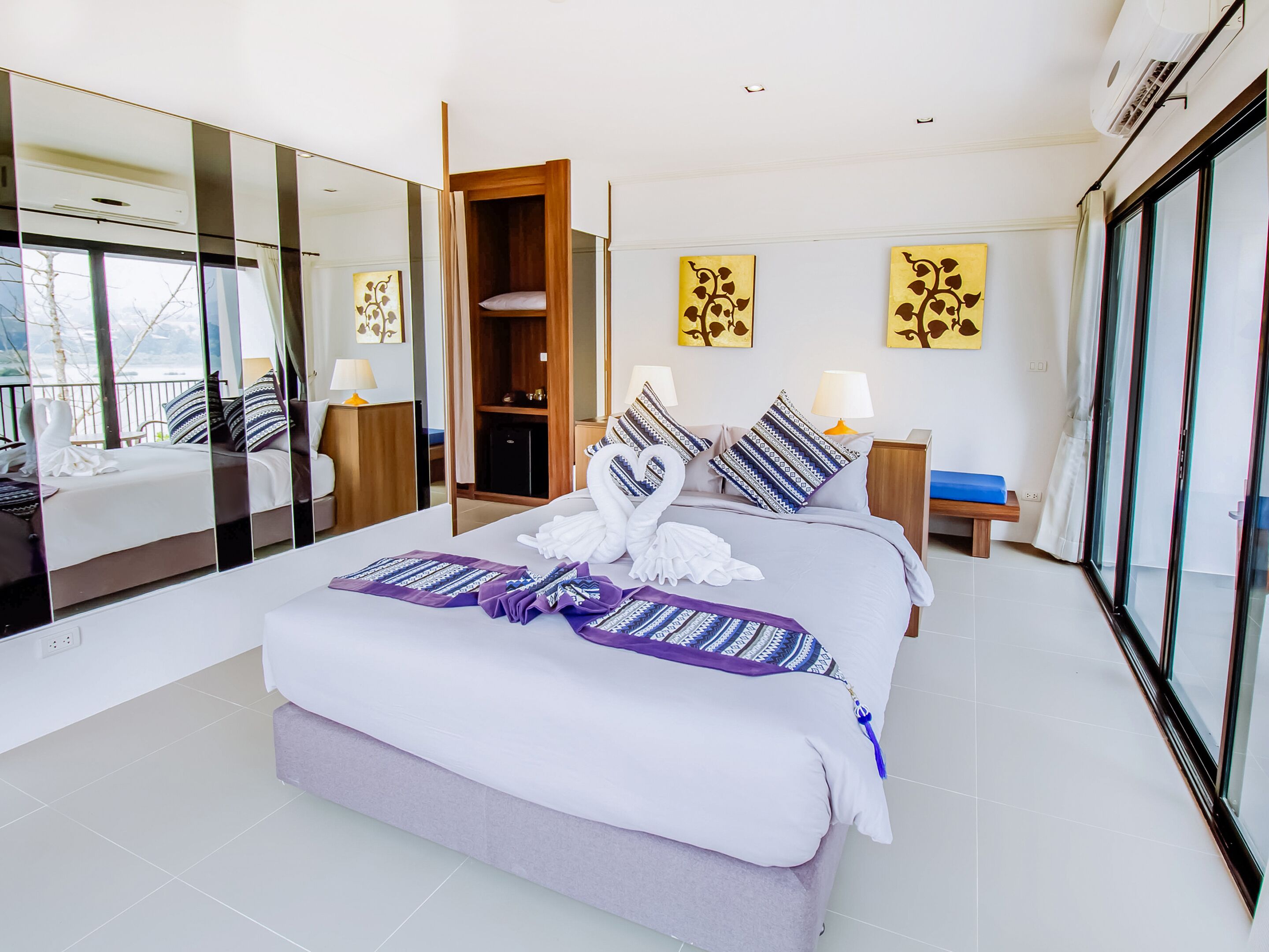 A modern hotel room with a large bed, a wooden wardrobe, a bedside table with a lamp, and a large window offering a view of the outdoors.