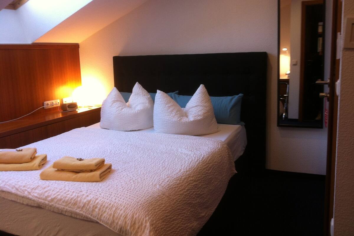 A neatly made bed with white pillows and folded towels, a bedside lamp, and a mirror on the wall.