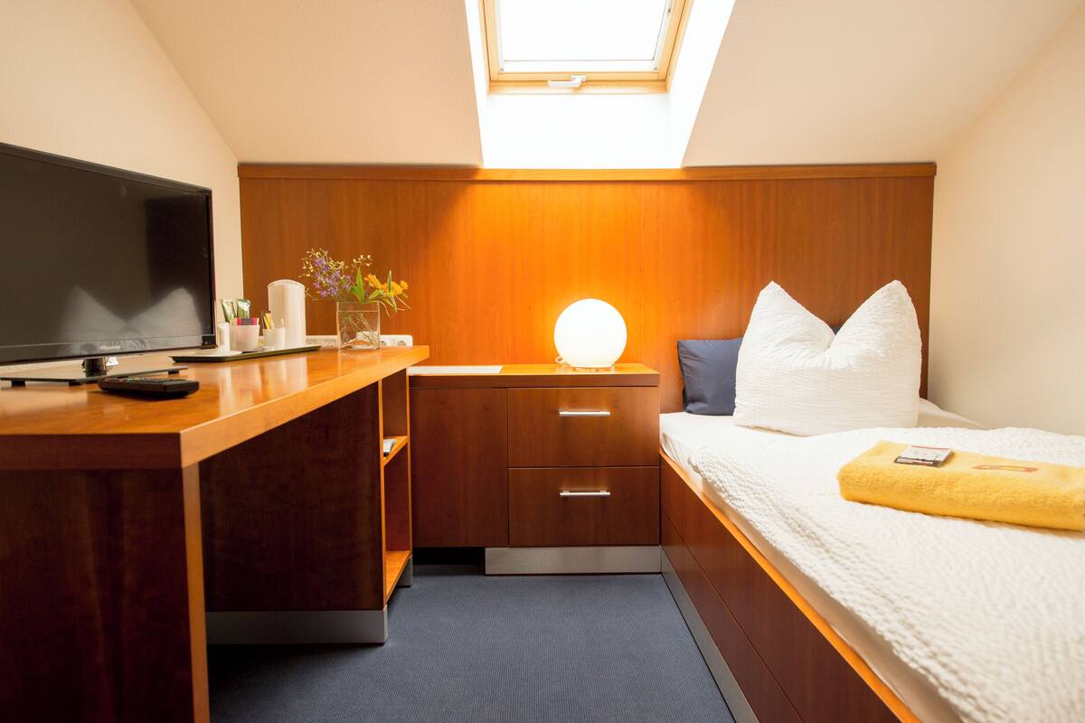 Habitación de hotel compacta con cama, mesita de noche, televisor y tragaluz.