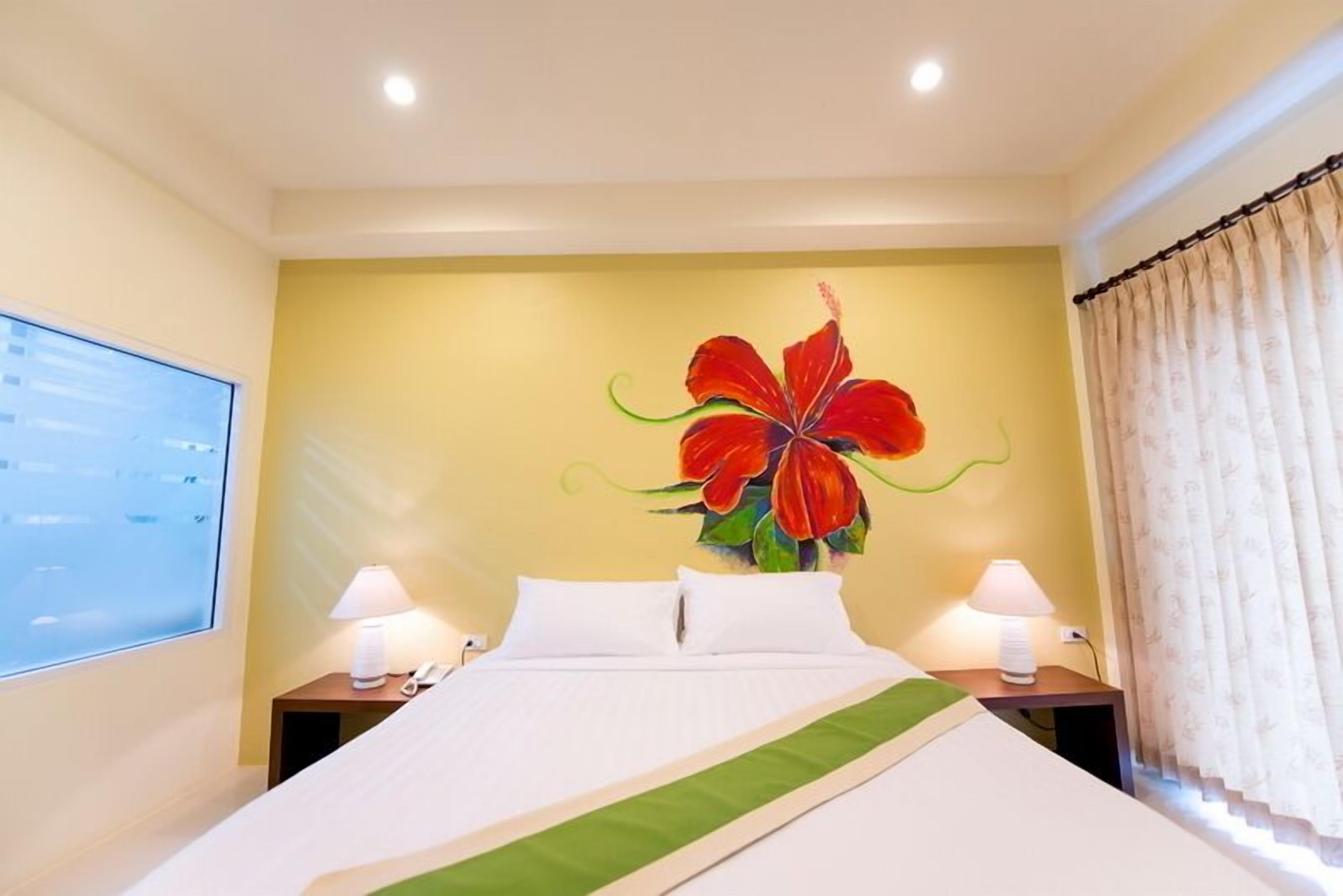 A hotel room with a bed, two bedside tables with lamps, a large floral artwork on the wall, and a window with curtains.