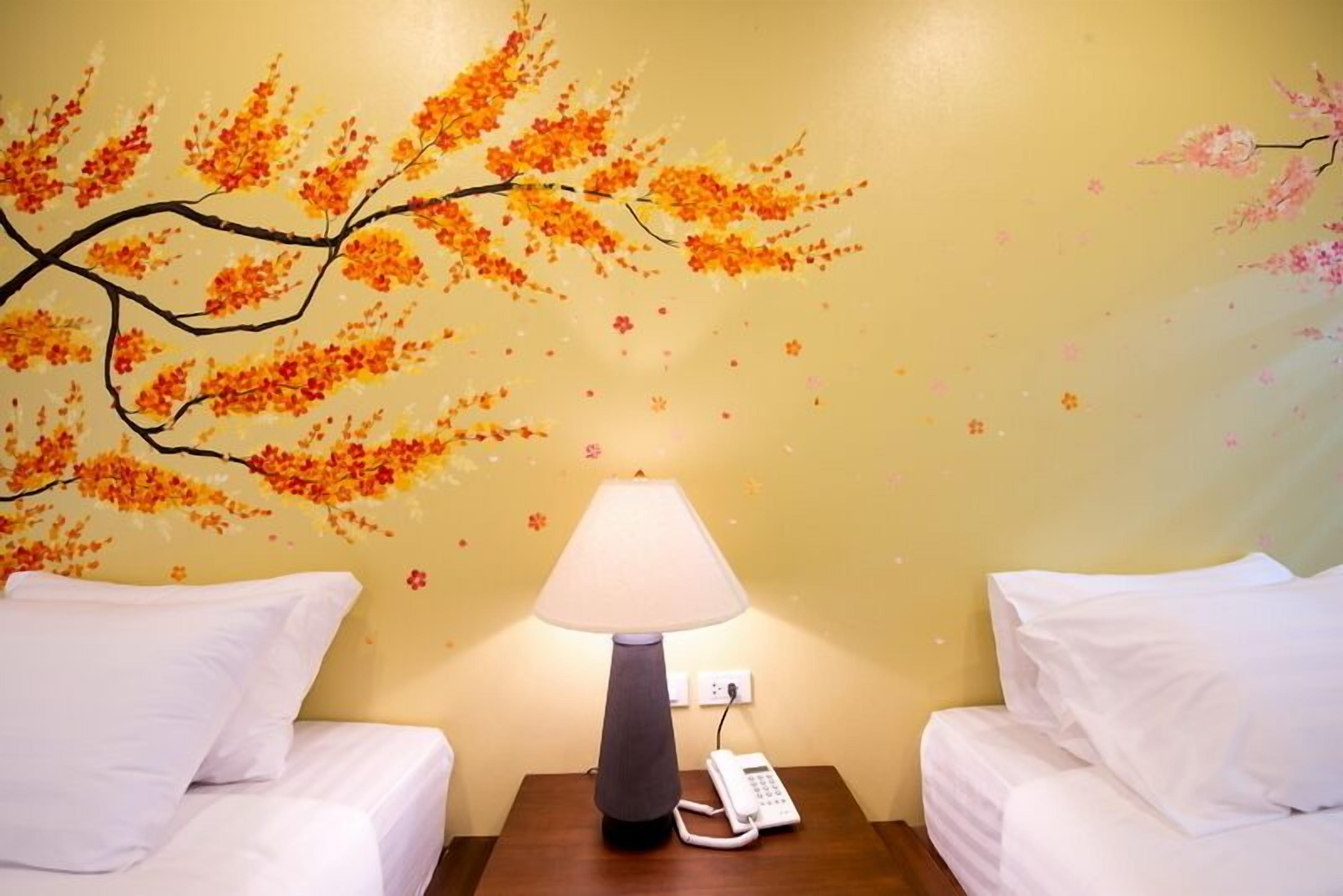 Habitación de hotel con dos camas, una mesita de noche con lámpara y un mural en la pared de hojas otoñales.