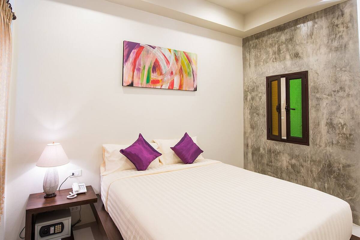 A hotel room with a bed, a nightstand with a lamp and phone, a painting on the wall, and a door with a green screen.