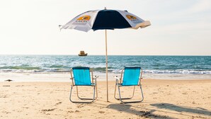 Two beach chairs under a beach umbrella with a logo, facing the ocean. - Hotel Ilhasul (Florianópolis)