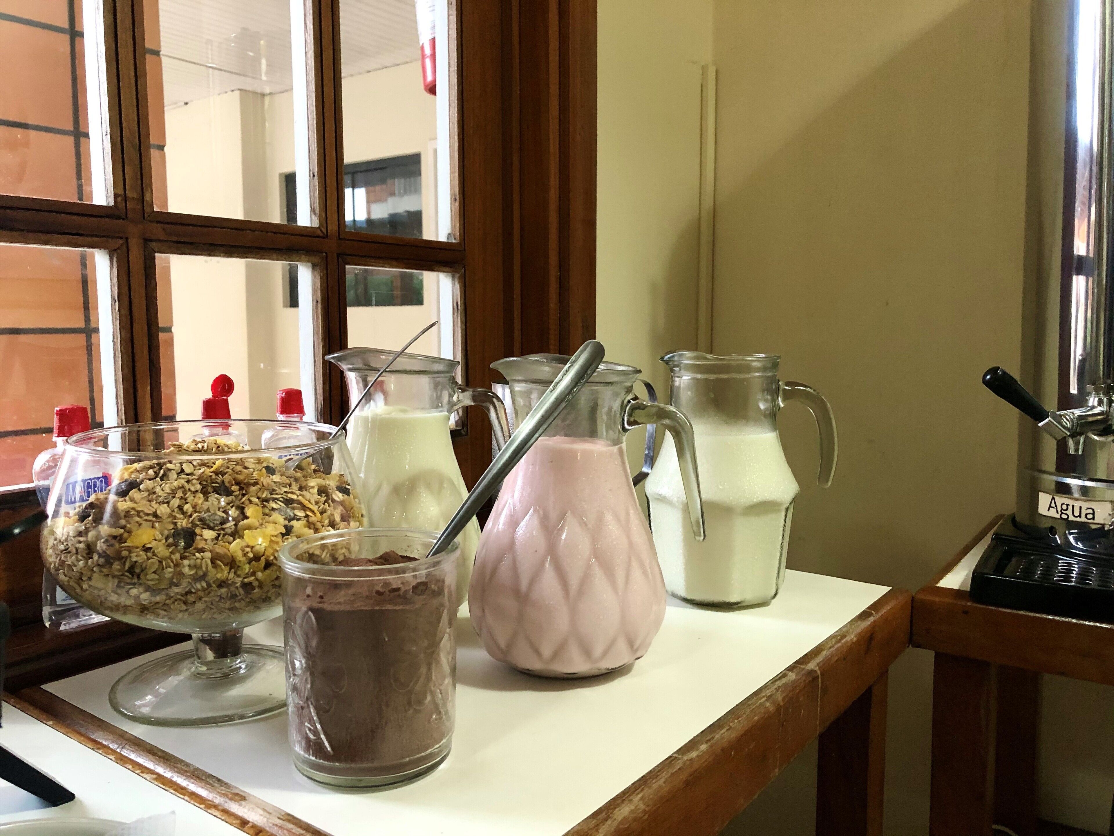 Un buffet pour le petit-déjeuner proposant divers aliments et boissons, notamment un bol de granola, une carafe de lait, un verre de lait au chocolat et une carafe d’eau.