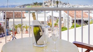 Un balcon avec une table dressée pour deux personnes, une bouteille de champagne et deux verres. En arrière-plan, on aperçoit un port de plaisance avec des bateaux et une ville côtière.