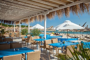An outdoor dining area with blue tables and wicker chairs, overlooking a beach with umbrellas and people. - Saint Thomas Holiday Village (Sozopol)