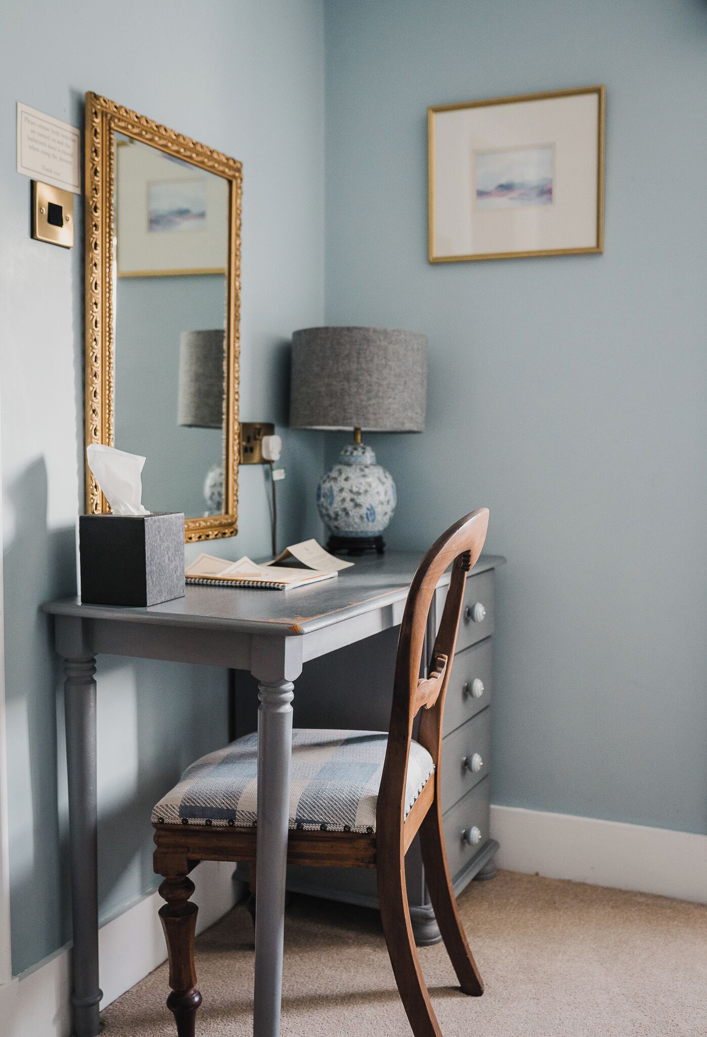 A study room with a desk, chair, lamp, and framed artwork on the wall.