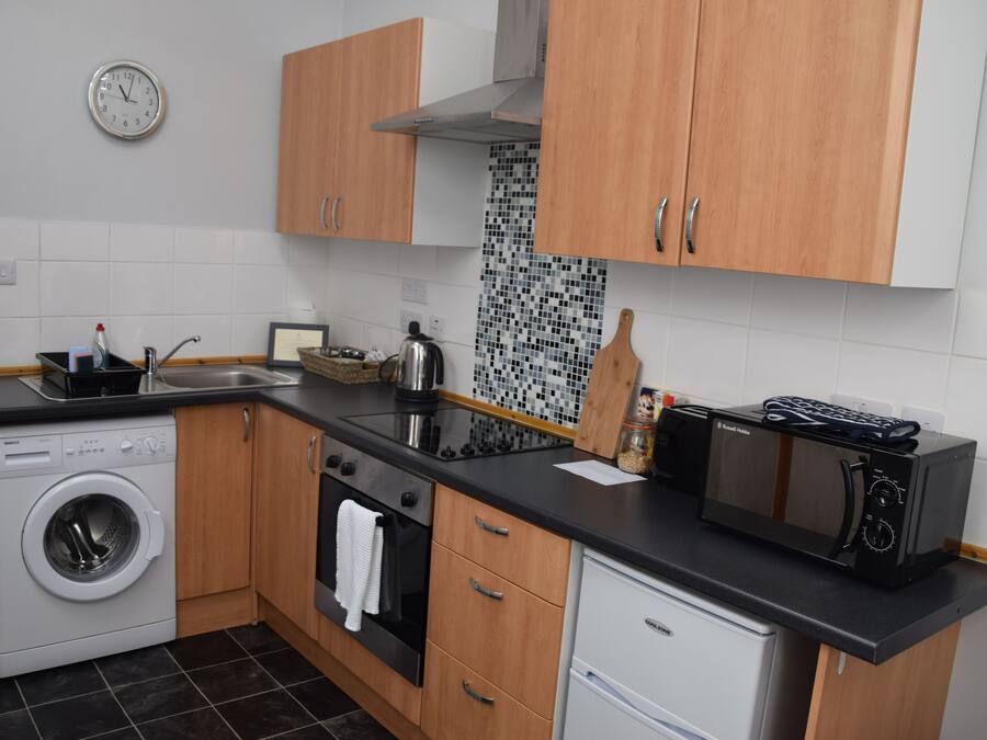 A compact kitchen with wooden cabinets, a washing machine, a microwave, and a kettle.